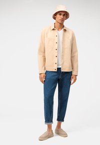 Giacca di cotone beige con chiusura a bottoni, t-shirt grigia chiara, pantaloni in denim blu e scarpe slip-on beige. Include anche un cappello a secchiello beige.