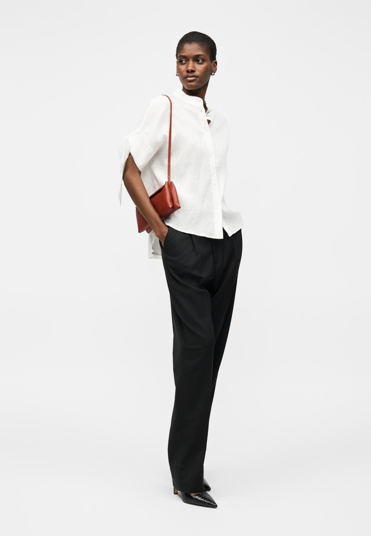 Femme portant un chemisier blanc à manches courtes, un pantalon noir taille haute, des talons pointus noirs et un sac à main rouge, debout les mains dans les poches.