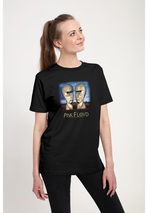 Kvinde med brunt hestehale iført sort Pink Floyd T-shirt med to metalliske hoveder vendt mod hinanden med en blå himmel i baggrunden.