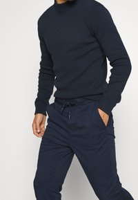 Maglione a maglia navy con colletto alto e polsini a coste abbinato a pantaloni navy con cordoncino. Entrambi gli articoli presentano una texture liscia e una vestibilità rilassata.