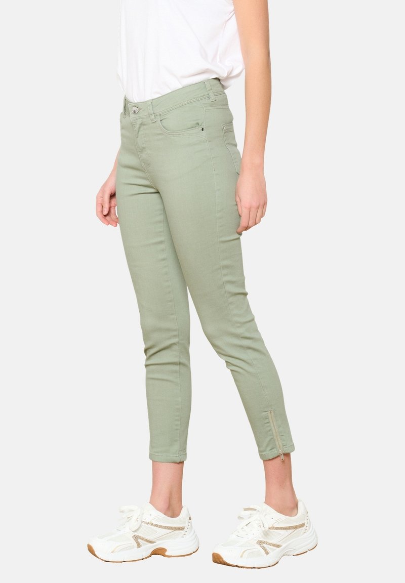 Heledad värve puuvillased skinny teksad, millel on lukuga sääredetail, kombineerituna valgete sneakersitega, millel on kuldsed aksessuaari.