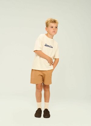 Krémové bavlnené tričko s námorníckym nápisom "Summer Crew", doplnené o pieskové šortky. Model má na sebe krémové ponožky a tmavohnedé slip-on topánky.