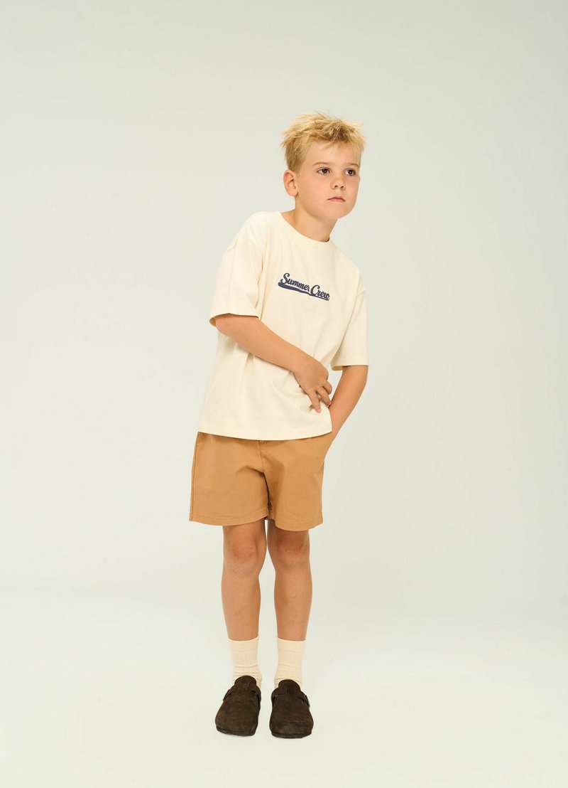 Camiseta de algodón color crema con texto en azul marino "Summer Crew", combinada con pantalones cortos color caqui. El modelo lleva puestos calcetines color crema y zapatos oscuros de deslizar.