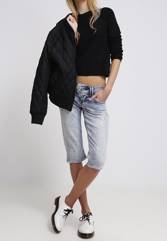 Femme portant un pull noir court, un jean bleu clair au genou, des chaussures blanches à lacets, tenant une veste matelassée noire sur l'épaule.