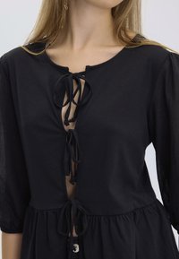 Robe noire avec un fermé à nouer à l'avant, des manches trois-quarts et un ourlet évasé. Fabriquée en tissu léger au toucher lisse.