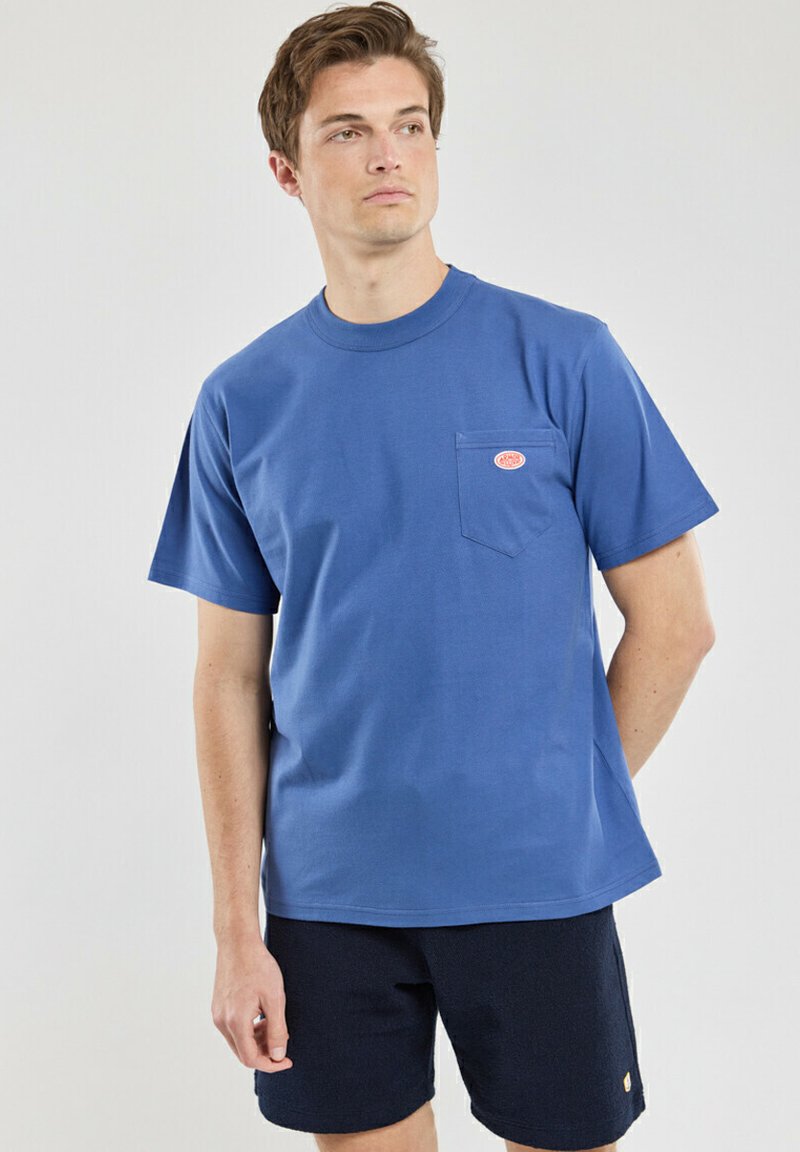 Blaues Baumwoll-T-Shirt mit Rundhalsausschnitt, kurzen Ärmeln und einer linken Brusttasche mit einem kleinen orangenen Logo. Getragen mit dunklen Shorts.