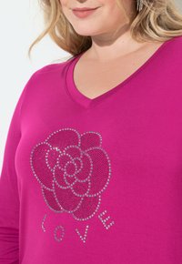 Maglietta fucsia a maniche lunghe con scollatura a V e un design di una rosa realizzata in strass con la parola "AMORE" sotto di essa.