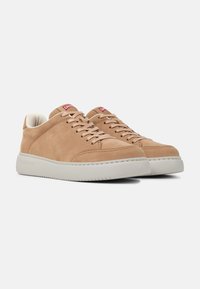 Light brown suede sneakers with white rubber soles, minimal design, flat laces, and red logo tag on the tongue. Soft, smooth texture.