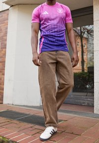 Mężczyzna ubrany w fioletową i różową koszulkę sportową Adidas, luźne brązowe spodnie i białe trampki, idący po ceglanej nawierzchni w pobliżu wejścia do budynku.