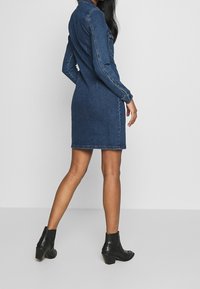 Jeanskjolsklänning i mörkblått, med lång ärm, knäppning framtill och en figurnära silhuett, bärs med svarta ankelstövlar.