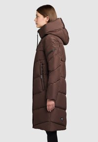 Ruskea toppatakki, jossa korkea kaulus ja huppu, vaakasuorat tikatut osiot, vetoketjulliset sivutaskut ja logokyltti helmassa.