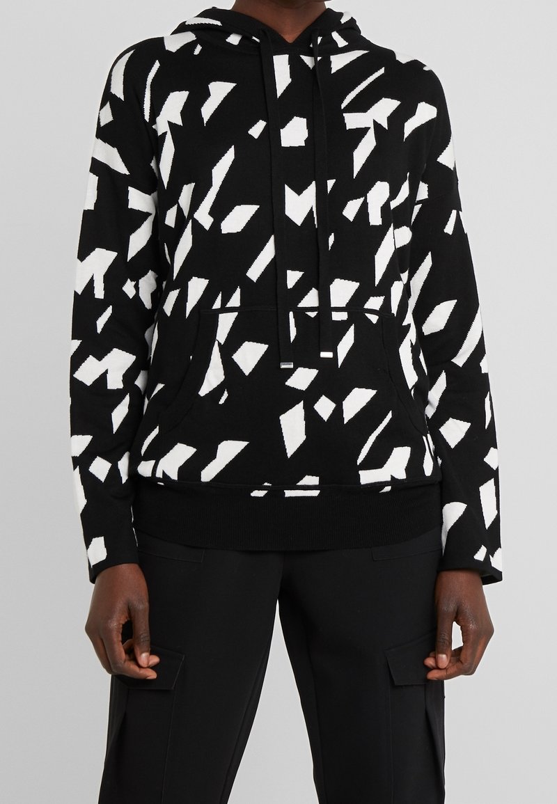 Sweat à capuche noir avec des formes géométriques abstraites blanches, poche avant, cordons de serrage, porté avec un pantalon noir doté de poches cargo latérales.