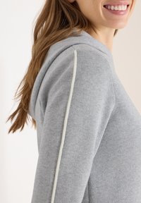 Uśmiechnięta osoba nosząca jasnoszarą dzianinową bluzę z kapturem z białym paskiem wzdłuż rękawa, widziana z profilu od ramion w górę.