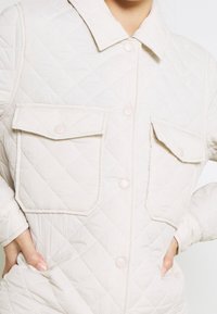 Kurtka pikowana w jasnobeżowym kolorze z fakturowanym wzorem diamentowym, wyposażona w dwie przednie kieszenie oraz zapięcia na guziki z przodu i na mankietach.