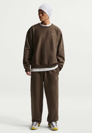 Mann iført brun Nike-genser og bukser, hvit lue, og grå joggesko med gule såler, stående med én hånd i lommen.