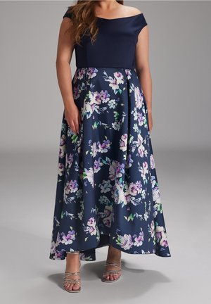 Kobieta w granatowej sukni z odkrytymi ramionami i spódnicą w fioletowo-białe kwiaty, ubrana w srebrne sandały na paski, stoi na tle jednolitego szarego tła.