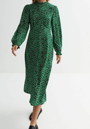 Femme marchant dans une robe midi verte à manches longues et col montant avec un motif abstrait noir et beige et des sandales noires.