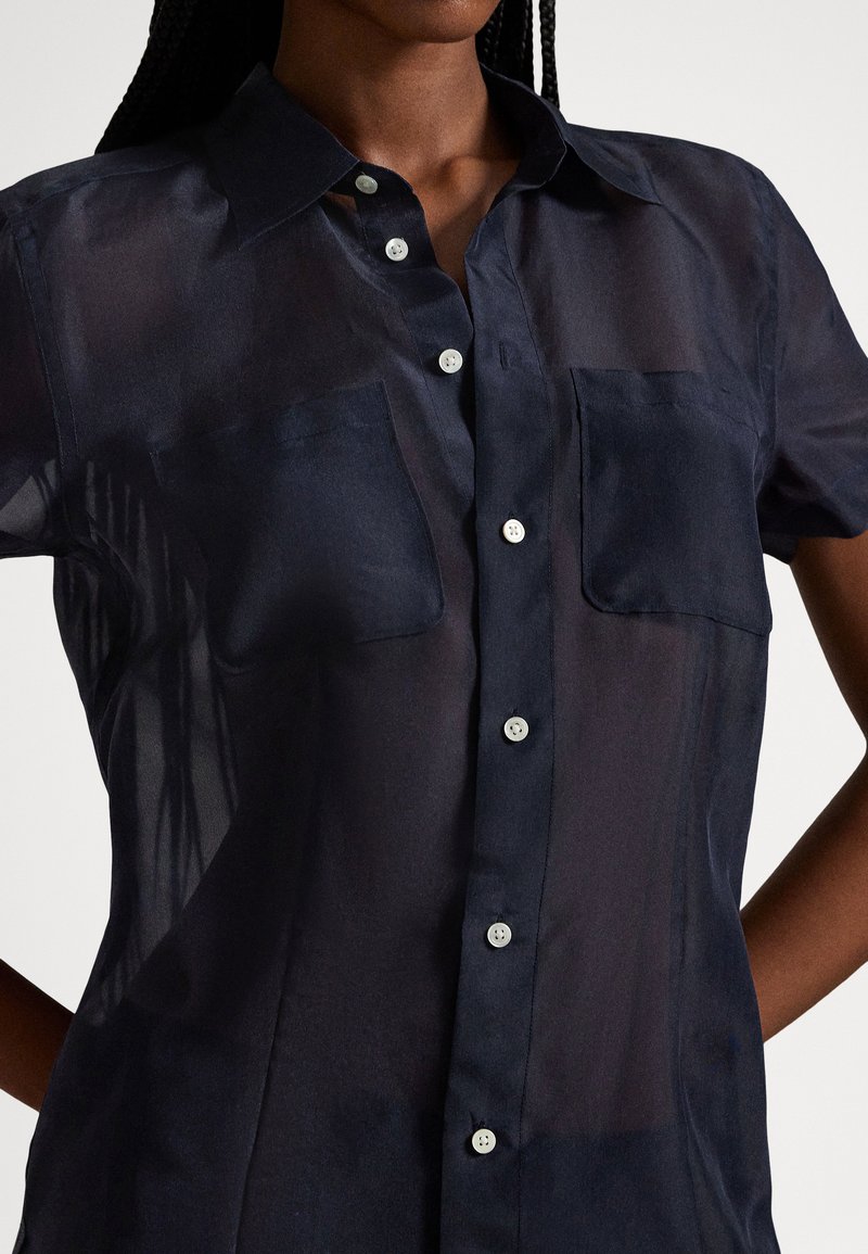 Frau trägt eine dunkle, transparente kurzärmelige Hemdbluse mit Brusttaschen, steht mit den Händen in der Hüfte vor einem schlichten Hintergrund.