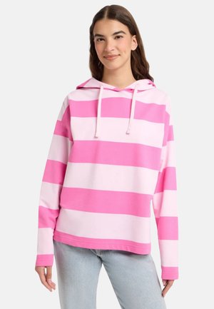 Young woman wearing a loose pink and white wide striped hoodie and light blue jeans, standing against a plain background.