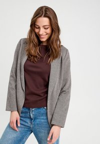 Blazer en pied-de-poule grise et noire avec une coupe ajustée, des revers crantés et des manches longues, superposé sur une chemise marron et un jean bleu.