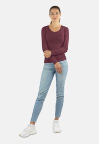 Langärmliges, kastanienrotes Oberteil, kombiniert mit hellblauen Slim-Fit-Jeans und weißen Sneakern. Das Oberteil hat einen runden Ausschnitt und eine taillierte Passform.