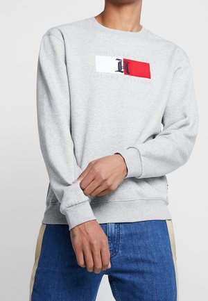 Mann trägt hellgrauen Pullover mit weißem und rotem Flaggenaufnäher und dunkelblaue Jeans, zieht mit einer Hand den Ärmel hoch.