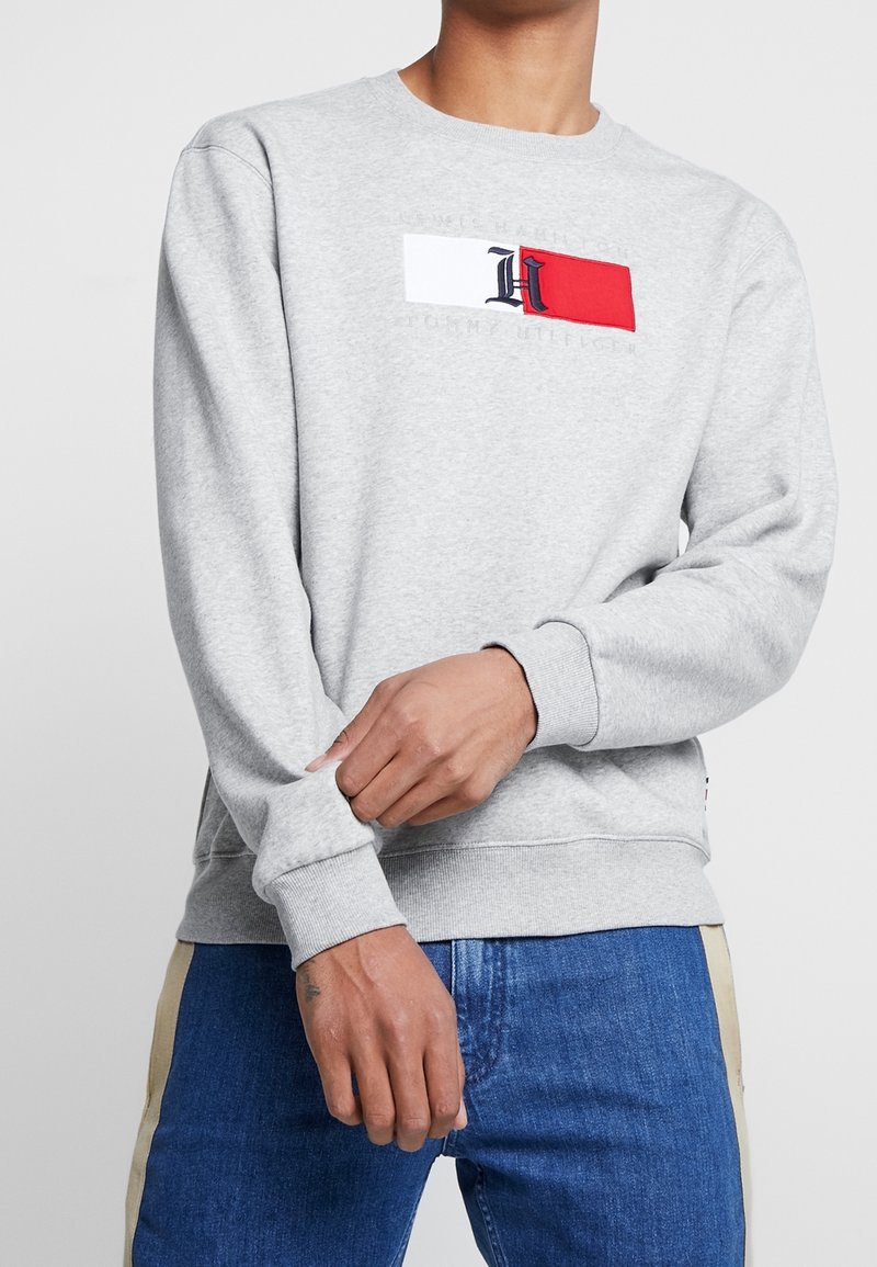 Homme portant un sweat-shirt gris clair avec un écusson de drapeau blanc et rouge et un jean bleu foncé, ajustant la manche avec une main.