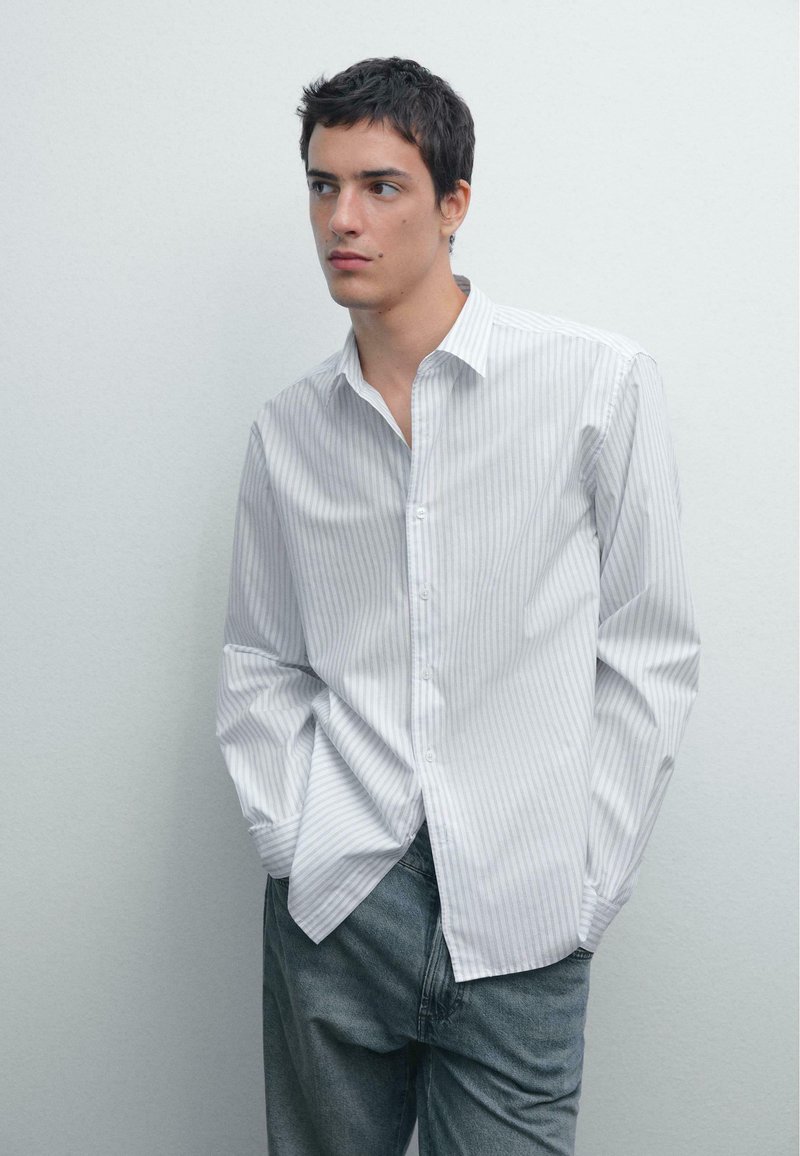 Camicia bianca con strisce verticali blu chiaro, maniche lunghe e colletto classico, abbinata a pantaloni in denim grigi.