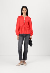 Blusa rossa a maniche lunghe con design a chiave e legature al colletto; vestibilità ampia, abbinata a jeans grigio scuro e scarpe basse beige.