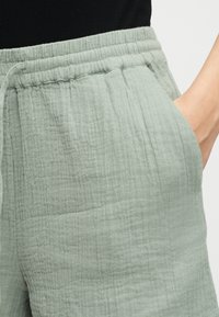 Personne portant un pantalon vert clair texturé avec une ceinture élastique et la main placée dans la poche droite.