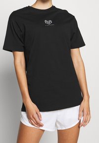 Černé bavlněné tričko, krátké rukávy, kulatý výstřih, s bílým geometrickým logem na hrudi, nošeno s bílými šortkami.