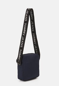 Niebieska torebka crossbody z tkaniny z czarnym paskiem, na którym widnieje napis "TOMMY JEANS" w kolorze białym. Zamek błyskawiczny oraz regulowany pasek.