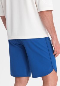 Modré sportovní šortky vyrobené z prodyšné tkaniny, s elastickým pasem a bočními rozparky pro pohyblivost. Jednoduchý design bez vzorů.