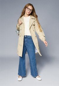 Beige vandtæt trench coat, blå brede bukser, hvide flade sko og runde briller. Enkel design med knapper og en glat tekstur.