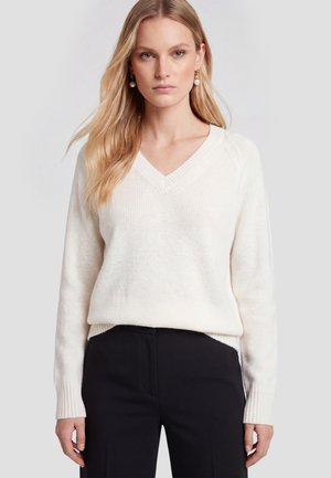 Kvinde med langt blondt hår iført hvid V-hals sweater og sorte bukser, set forfra mod en ensfarvet lys baggrund.