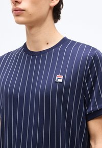 Νεαρό άτομο που φοράει μπλούζα σε ναυτικό μπλε χρώμα με λευκές κάθετες ρίγες και ένα μικρό κόκκινο, λευκό και μπλε λογότυπο Fila στο στήθος.