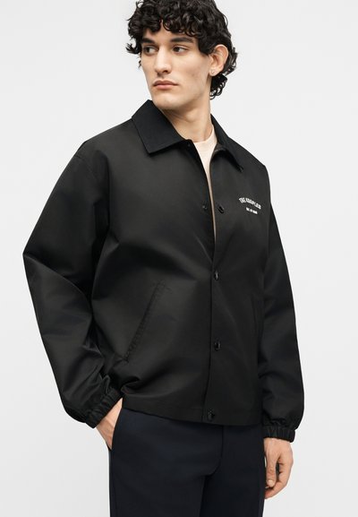 Νέος άνδρας με σγουρά μαλλιά που φορά μαύρο σακάκι με κουμπιά, ελαστικές μανσέτες και λευκό κείμενο στο αριστερό στήθος, κοιτάζοντας προς το πλάι.