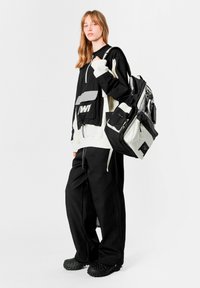 Chaqueta negra y blanca con cremallera y bolsillo delantero, combinada con pantalones anchos negros de pierna ancha y textura, y zuecos negros. Mochila grande con múltiples bolsillos.