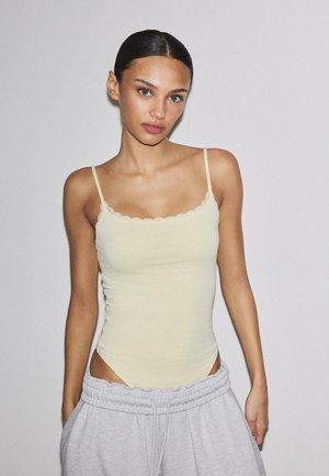 Kvinde med glat tilbagefejet hår iført en lysegul camisole med blonder og lysegrå joggingbukser mod en ensfarvet grå baggrund.