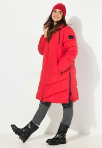 Cappotto imbottito rosso con un design lungo e trapuntato, dotato di tasche laterali e chiusura con zip. Indossato con jeans grigi e stivaletti neri.