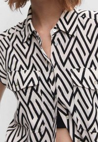 Chemise à manches courtes en tissu léger, avec un motif zigzag noir et blanc, deux poches poitrine à bouton et un col.