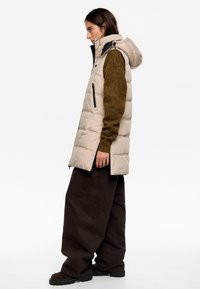 Gilet imbottito beige con cappuccio, dotato di imbottitura spessa, tasche con zip nere e vestibilità over size, abbinato a un maglione verde scuro e pantaloni marroni.