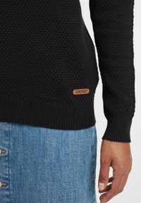 Schwarzer, strukturierter Pullover mit geripptem Saum und Bündchen, der ein Logo-Patch aus beige Leder an der Seite trägt. Kombiniert mit einem blauen Jeansrock.