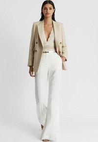 Beige dubbelknäppt blazer över en spetsblus, kombinerad med vita högmidjade byxor med utsvängd ben, som har sidsömmar och ett smalt bälte.
