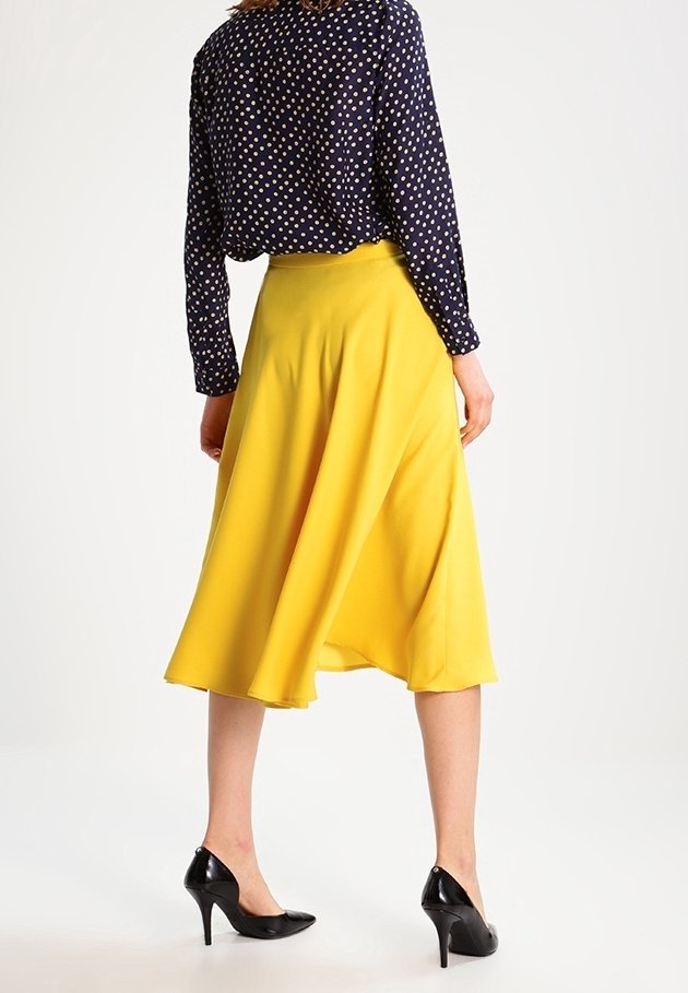 Femme portant un chemisier bleu marine à pois, une jupe midi jaune et des talons hauts noirs, debout de dos sur un fond blanc.