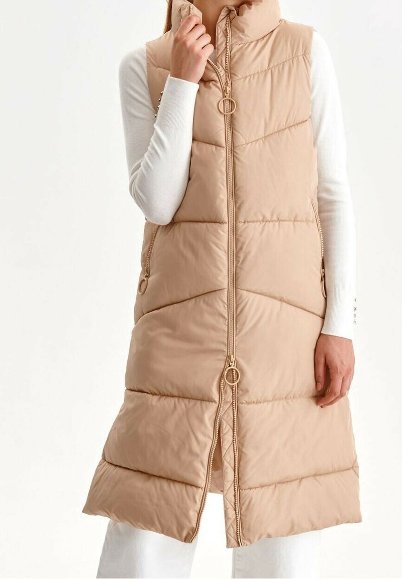 Beige Steppweste mit hochgeschlossenem Kragen, vertikal gestepptem Design, Reißverschluss vorne und goldenen Hardware-Elementen. Verfügt über Seitentaschen.