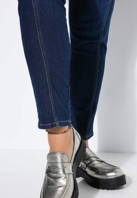 Metalické stříbrné mokasíny s robustní černou podrážkou, nošené s tmavě modrými přiléhavými džíny s ohrnutým lemem a viditelnými zvětšenými švy.