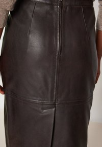 Jupe en cuir marron avec un design ajusté, fermeture éclair  à l'arrière et détails de couture subtils. La texture est lisse avec une finition élégante.