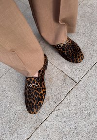 Luipaardprint loafers van textiel met gouden hardware-details, gecombineerd met tan broek, rustend op een stenen oppervlak.