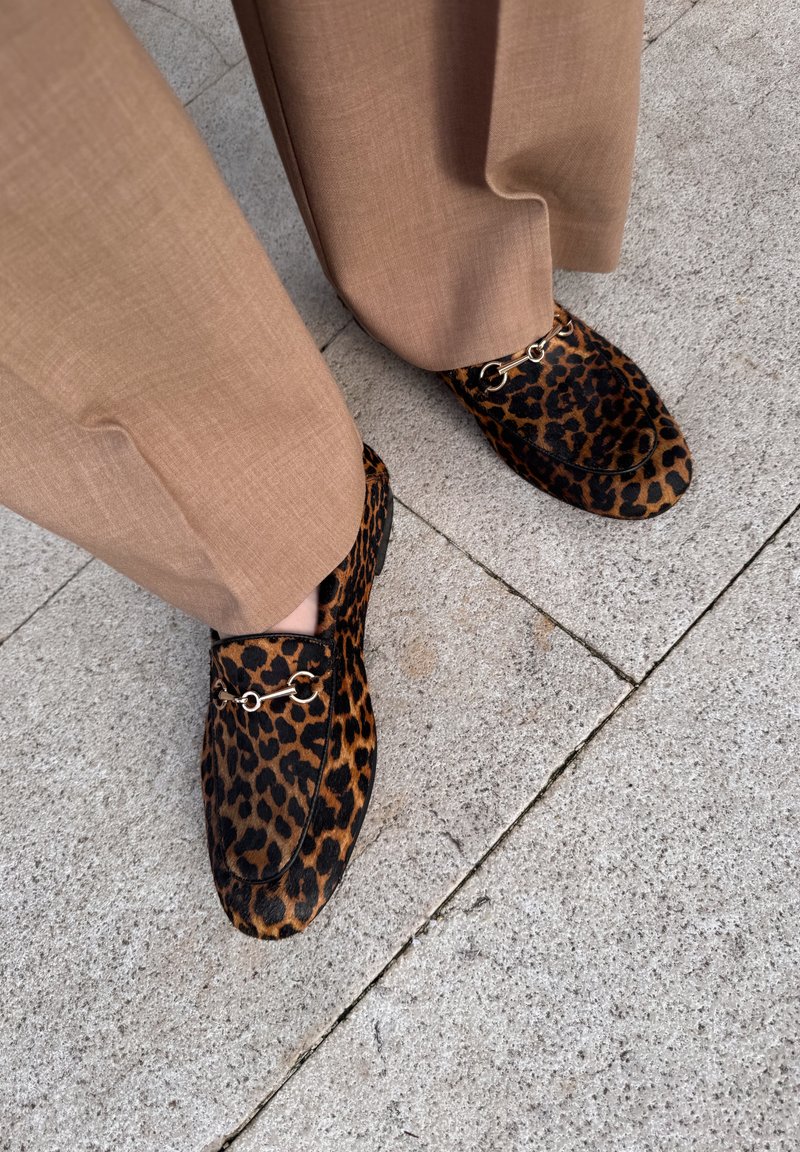 Luipaardprint loafers van textiel met gouden hardware-details, gecombineerd met tan broek, rustend op een stenen oppervlak.